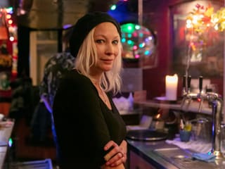 Eine Frau mit blonden Haaren, schwarzer Wollmütze und schwarzem Shirt steht hinter einem Bartresen. Eine Frau mit blonden Haaren, schwarzer Wollmütze und schwarzem Shirt steht hinter einem Bartresen.