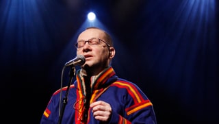 Wimme in traditioneller blauer Samen-Kleidung steht mit geschlossenen Augen vor einem Mikrofon.