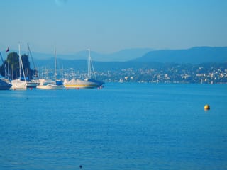 Am Ufer des See liegen grössere Segelschiffe und Moterboote vor Anker, im Hintergrund die Stadt, darüber klarerer hellblauer Himmel.