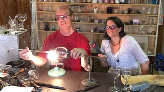 Ein Mann bearbeitet über einer Flamme Glas, daneben sitzt eine Frau mit SRF-Mikrofon. Beide tragen eine Schutzbrille. Ein Mann bearbeitet über einer Flamme Glas, daneben sitzt eine Frau mit SRF-Mikrofon. Beide tragen eine Schutzbrille.