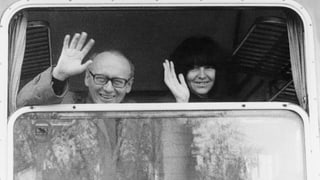Ernst Jandl und Friederike Mayröcker blicken lachend aus einem geöffneten Zugfenster und winken.