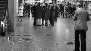 Schwarz-Weiss-Aufnahme: Tatort-Szene in einem Shopping-Center. Im Hintergrund stehen Schaulustige. 