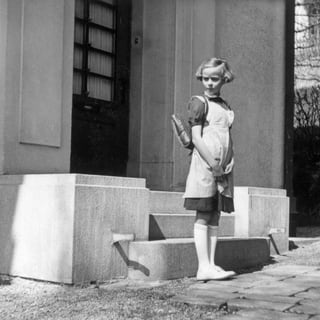 Ein Schwarz-weiss-Bild eines Mädchens, das mit Schulthek vor einem Haus steht und kritisch dreinblickt.
