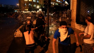 Einige Demonstraten, ein junger Mann mit einem weissen Tuch um Nase und Mund, hinten eine Polizeikolonne mit Schutzschildern.