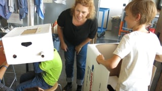 Zwei Kinder halten zwei grosse Schachteln. Sie sind daran, aus diesen Schachteln Zeitmaschinen zu bauen. Im Hintergrund steht die Gründerin Sabine Gysin, die sich die Maschinen erklären lässt.