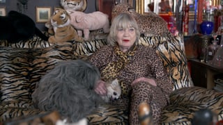 Eine Frau im Leopardenkostüm sitzt auf einem Sofa mit Zebramuster. Sie streichelt einen Hund. Auf dem Sofa liegen Stofftiere, darunter ein Tiger und ein Schwein. 