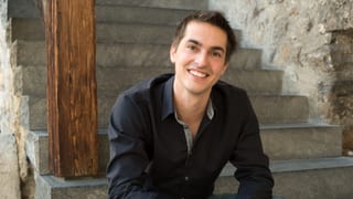 Ein junger Mann im schwarzen Hemd sitzt lächelnd auf einer Treppe.
