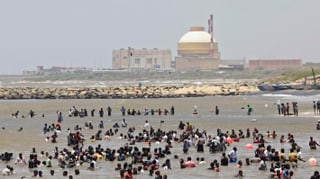 Menschen stehen an einem Strand im Wasser, im Hinterdrund ein AKW mit einer Betonkuppel.