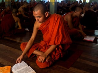 Ein buddhistischer Mönch sitzt im Schneidersitz und liest in einem Glaubenstext, im Hintergrund tun es ihm viele gleich.