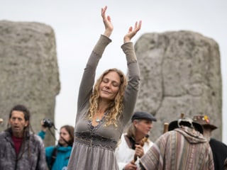 Frau steht mit erhobenen Händen und geschlossenen Augen vor Steinkreis