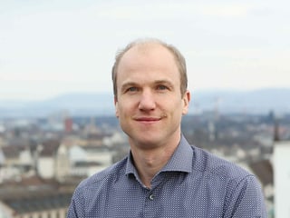 Ein Mann in einem blauen Hemd steht auf einem Balkon im freien. Hinter ihm sieht man Umrisse einer Stadt.