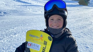 Gaël hält sein Snowboard in der Hand, steht auf der Piste und grinst in die Kamera