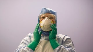 Person in einem Schutzanzug mit blauber Haube und grünen Gummihandschuhen.