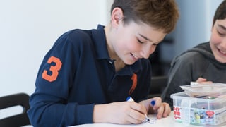 Ein Junge sitzt lachend an einem Tisch und zeichnet mit einem Buntstift auf einen Zettel.