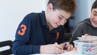 Ein Junge sitzt lachend an einem Tisch und zeichnet mit einem Buntstift auf einen Zettel. Ein Junge sitzt lachend an einem Tisch und zeichnet mit einem Buntstift auf einen Zettel.