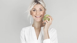 Eine blonde Frau hält sich einen grünen Apfel an die rechte Wange. 