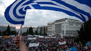 Ein Mann schwenkt eine griechische Fahne. Im HIntergrund Demonstrierende.