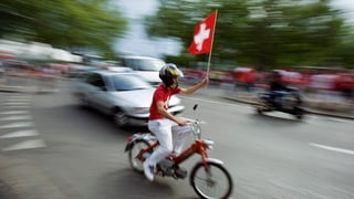Ein Schweizer Fussballfan fährt auf seinem Mofa durch die Strassen und hält dabei eine Schweizer Fahne in die Höhe.