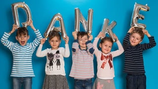 Fünf kleine Kinder mit den aufblasbaren Buchstaben D, A, N, K und E.
