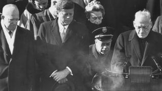 Feiner weisser Rauch steigt auf der linken Seite des Rednerpultes auf, Kennedy schaut interessiert
