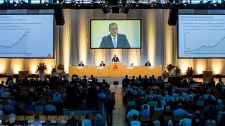 Versammlung in einem grossen Saal mit einem Bildschirm. Darauf Verwaltungsratspräsiden der Sika, Paul Hälg.