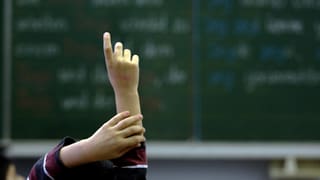 Ein Kind reckt die rechte Hand in die Höhe und meldet sich im Unterricht zu Wort. Im Hintergrund eine Wandtafel.