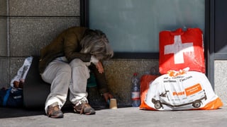 Ein obdachloser Mensch mit seinen Tüten sitzt vor einer Hausmauer.