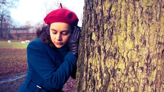Eine Frau mit Mütze und Mantel hält ihr Ohr an einen Baumstamm.