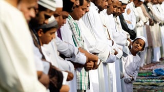Muslime stehen in einer Reihe, die Hände gefaltet, ein kleiner Junge ragt aus der Reieh und guckt sich um.