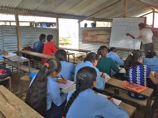 Blick in das Klassenzimmer. Der Lehrer schreibt auf eine weisse Tafel, die Kinder sitzen auf Holzbänken.