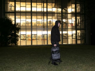 Mann in einem schwarzen Kapuzenpullover mit einem Rucksack steht vor einem Gebäude mit hell erleuchterter Glassfassade