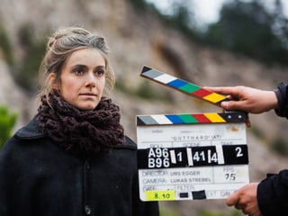 Eine junge Frau in dunklem Mantel steht vor einer geöffneten Filmklappe, die jemand ins Bild hält.