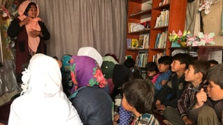 Jung Frau steht in einem Raum. Vor ihr sitzen Buben und Mädchen mit Kopftüchern. Jung Frau steht in einem Raum. Vor ihr sitzen Buben und Mädchen mit Kopftüchern.