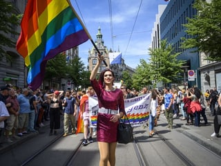 Eine Dragqueen oder Transfrau in einem glitzernden Kleid läuft an der Zürich Pride mit und schwingt dabei eine Regenbogenfahne.