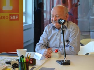 Ernst Gilgen sitzt vor dem Mikrofon und lacht verschmitzt vor sich hin.