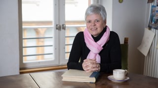 Eine Frau sitzt an einem Tisch. Ihre verschränkten Händen liegen auf einem Buch. 