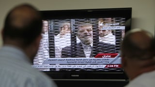 Zwei Männer schauen TV, auf dem Mursi hinter Gittern zu sehen ist