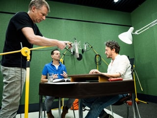 Drei Männer in einem Studio mit Mikrofonen und grünen Wänden.