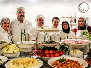 Eine Gruppe von Frauen in Kochuniform stehen hinter einem Tisch mit verschiedenen Speisen.