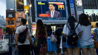Menschen stehen in einer städtischen Fussgängerzone vor einem grossen Bildschirm, auf dem Premier Abe spricht.