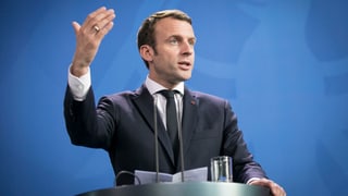 Macron vor blauem Hintergrund an einem Rednerpult, er gestikuliert.