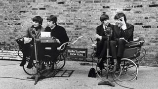 Vier Musiker sitzen auf zwei Kutschen auf einer Strasse.