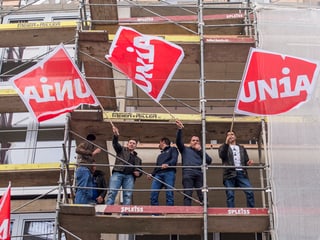 Bauarbeiter stehen auf Baugerüst und schwenken rot-weisse Unia-Fahnen.