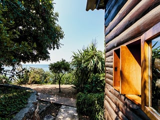 Hauswand aus Holz, daneben mediterraner Park und Sicht auf das Meer. 