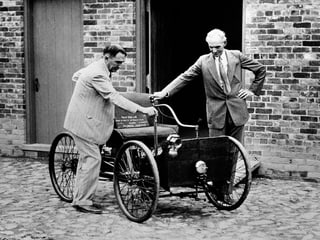 Ford (r.) mit dem ersten Auto, das er baute, auf einer Kopfsteinpflaster-Strasse vor einer Wand.