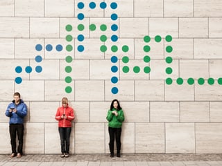 Drei junge Leute stehen an einer Wand und schauen jeweils auf ein Smartphone. Über ihren Köpfen sind gepunktete Linien zu sehen, die sie miteinander verbinden oder aus dem Bild hinausführen. 