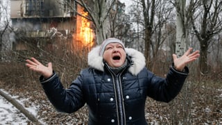 Eine Frau schreit ihre Frustration über die Zerstörung heraus, im Hintergrund brennt ein Haus.