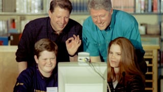 Zwei Männer stehen hinter zwei Schülern, welche an einem Computer sitzen.