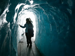 In einer Gletscherhöhle leuchtet das Eis bläulich, eine Frau berührt das Eis in der Höhle mit den Fingern, am Boden ist ein Steg aus Holz ausgelegt. 