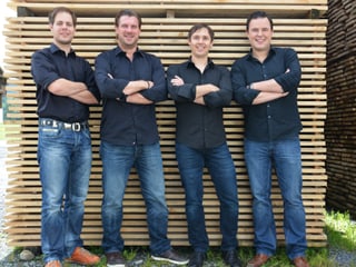 Die vier Musiker tragen Jeans und dunkelblaue Hemden und stehen vor einem Stapel Holzbrettern.